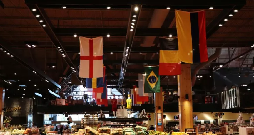 Fresh produce display inside a covered food market with several international flags hanging from the ceiling.